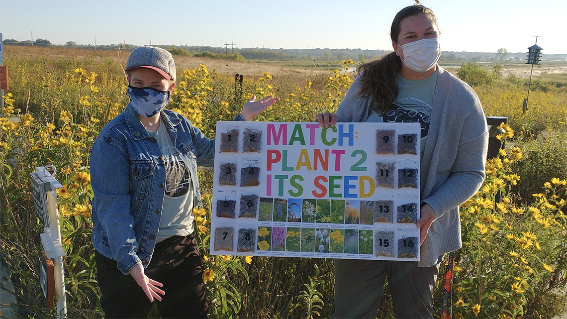"Two students holding poster about seed identification, with yellow flowers blooming in wetlands in background"